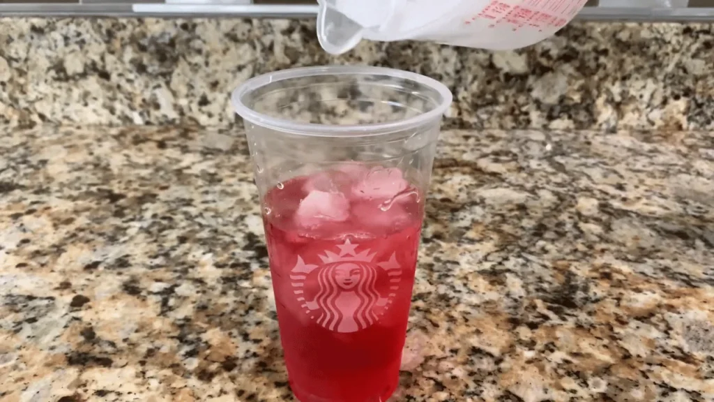 Ice cubes being added to a strawberry refresher drink in a clear cup for a homemade pink drink recipe