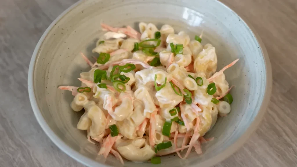 A close-up shot of a creamy Hawaiian macaroni salad mixed with shredded carrots and topped with sliced green onions in a grey ceramic bowl.