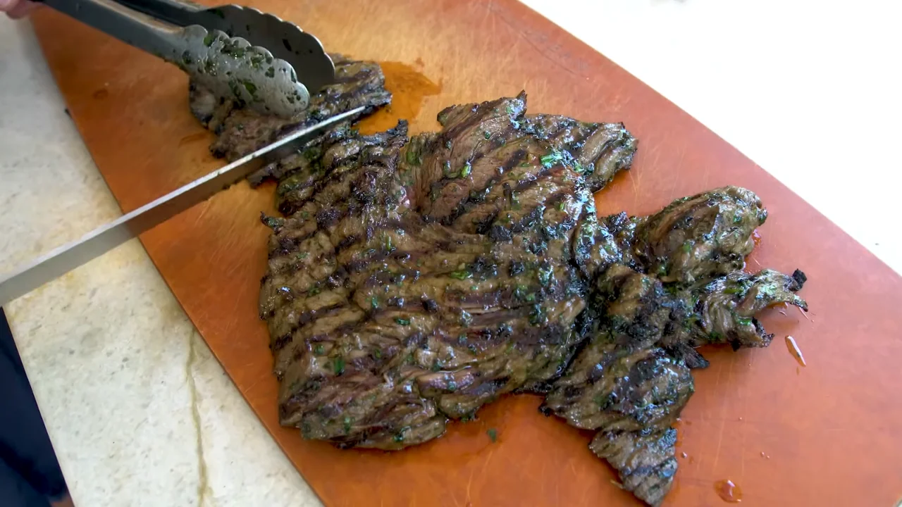 Grilled flank steak carne asada being sliced with a knife and tongs on a brown cutting board.
