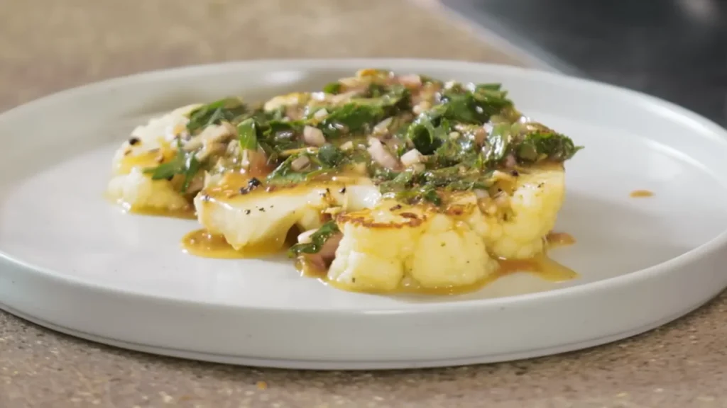 A thick grilled cauliflower steak served on a white plate, topped with a savory teriyaki glaze and freshly chopped green herbs and scallions.