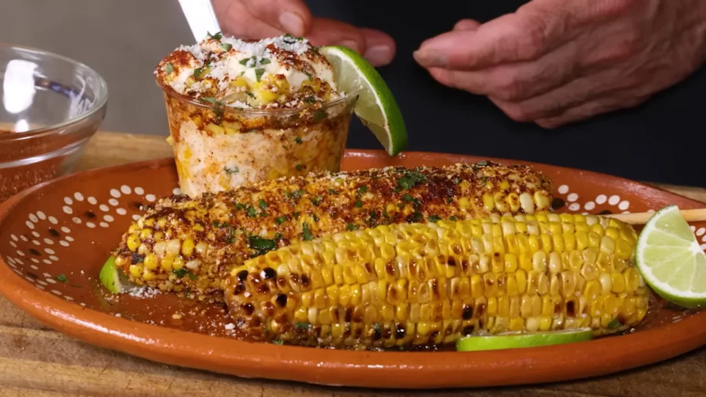 Two ears of grilled Mexican street corn on a brown plate, coated with mayonnaise, cheese, chili powder, and fresh cilantro, served with lime wedges and a small cup of corn.