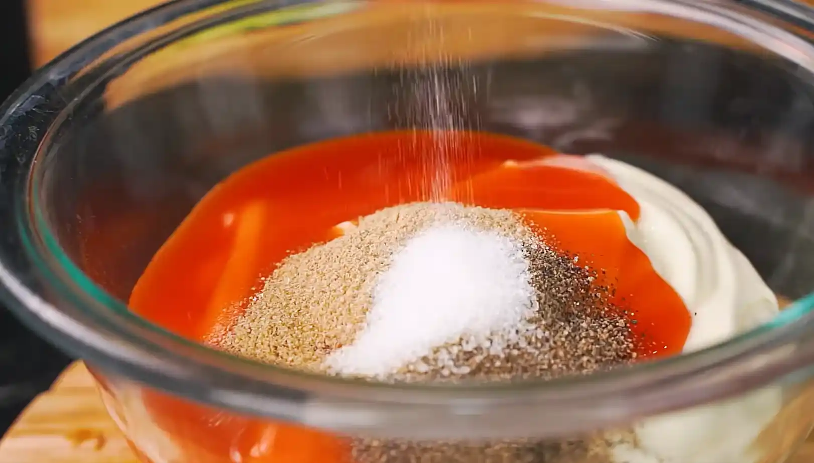 Hot sauce, mayonnaise, garlic powder, sugar, and black pepper being combined in a glass bowl for spicy mayo