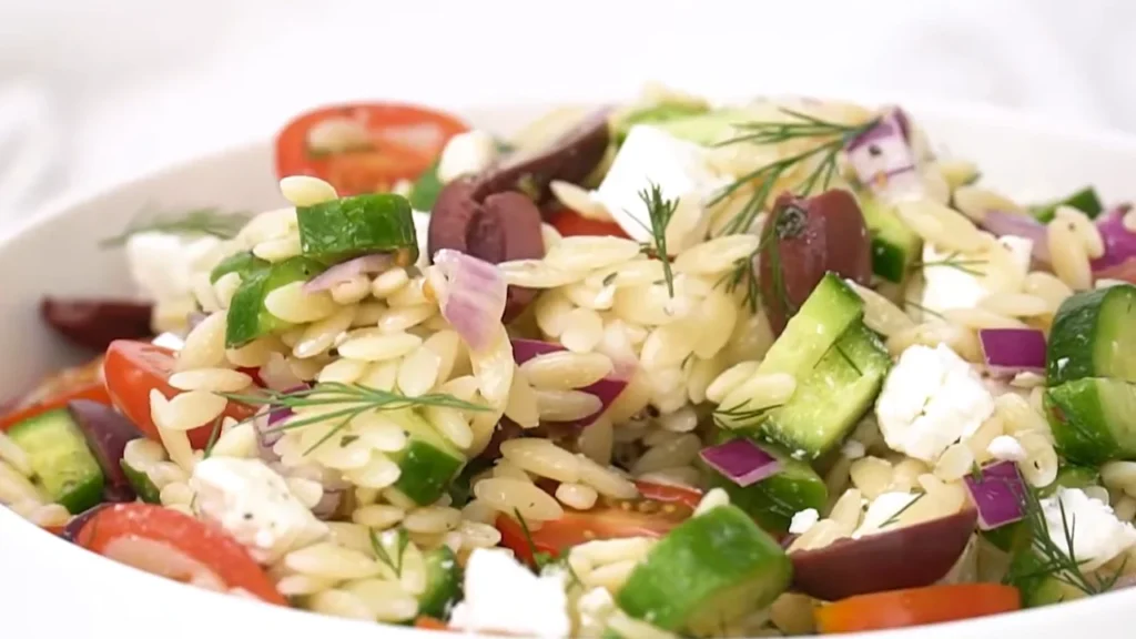 A close-up view of a Mediterranean orzo salad mixed with diced cucumbers, cherry tomatoes, kalamata olives, red onion, and crumbled feta cheese in a white bowl.