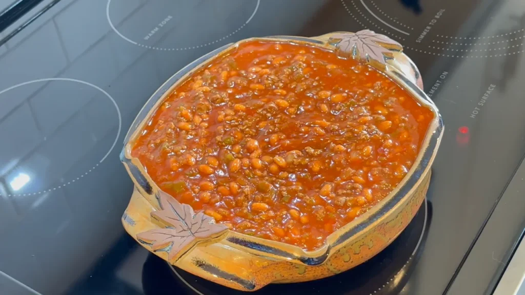 A top-down view of thick, bubbling sweet and savory baked beans mixed with bits of bacon and onions, cooking in a large skillet on a smooth black stovetop.