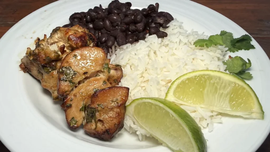 Grilled cilantro-lime chicken pieces served on a white plate with white rice, black beans, fresh cilantro leaves, and lime wedges.