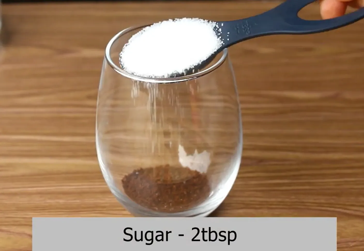 Pouring granulated white sugar over instant coffee in a clear glass.