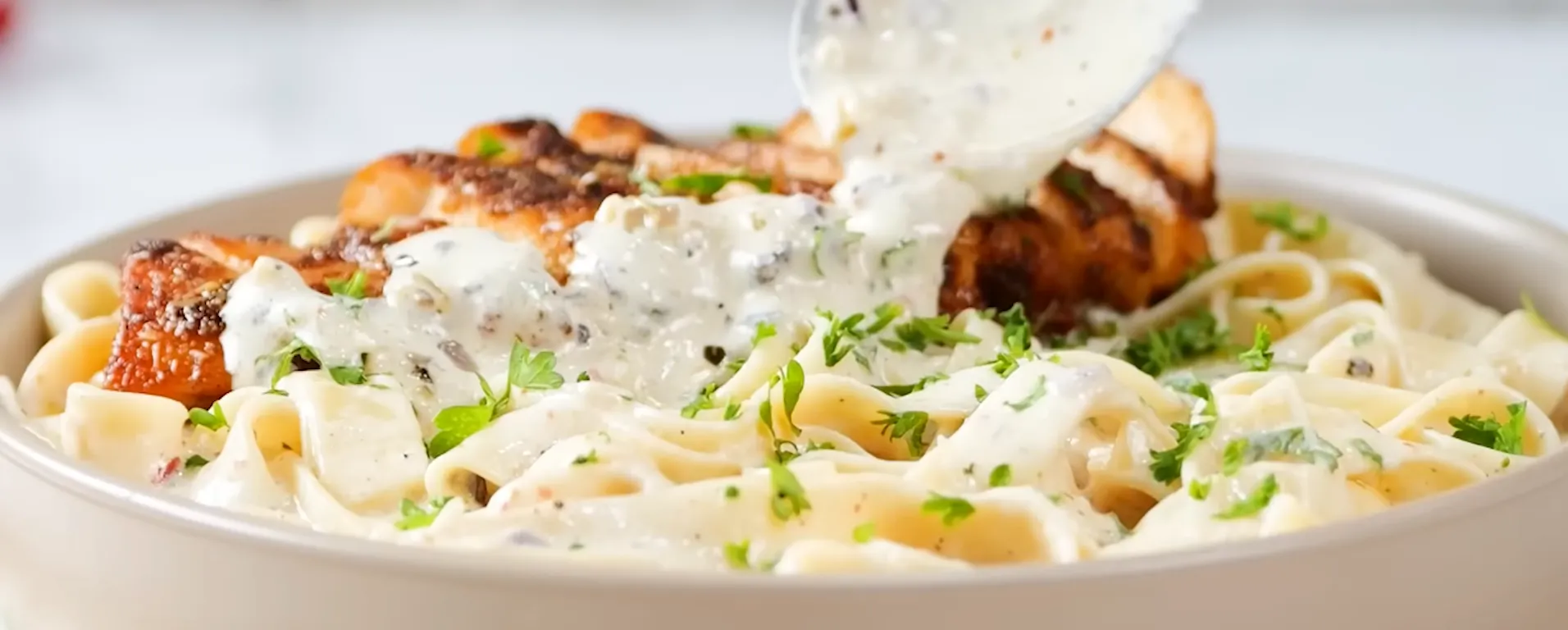 Close-up of creamy white Alfredo sauce being poured from a spoon onto chicken fettuccine pasta.