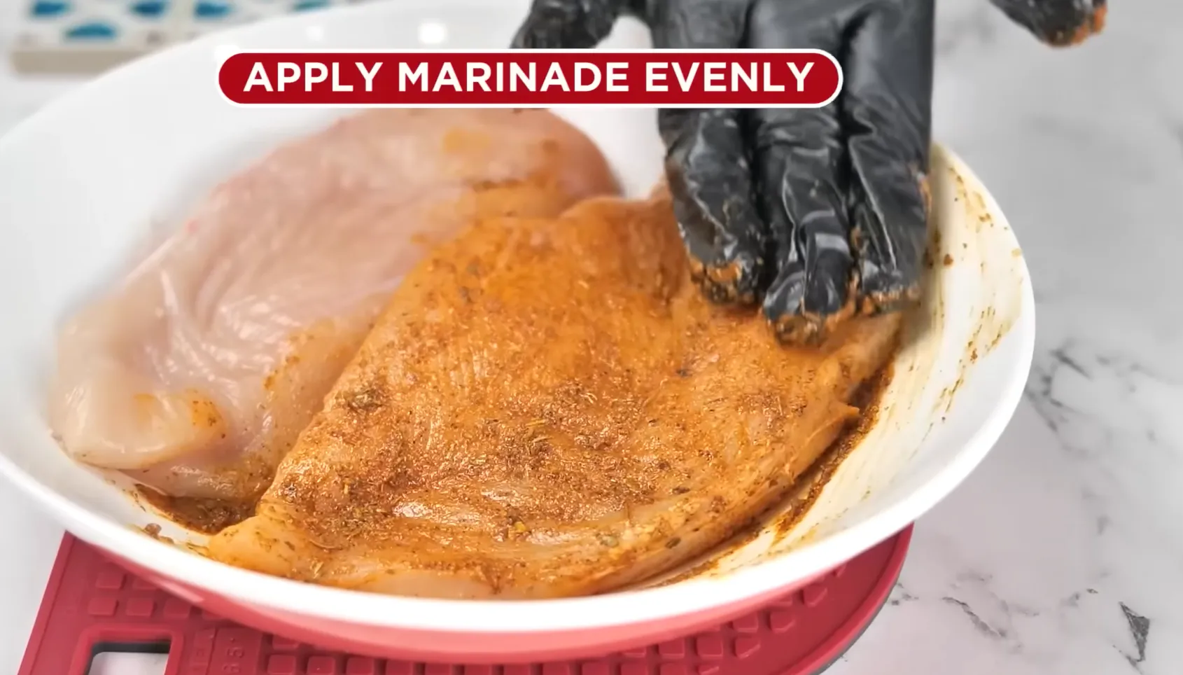 Applying orange spice marinade evenly on raw chicken breasts in a white bowl using black gloves.