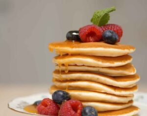 A tall stack of fluffy homemade golden-brown American pancakes topped with a melting butter cube and dripping maple syrup