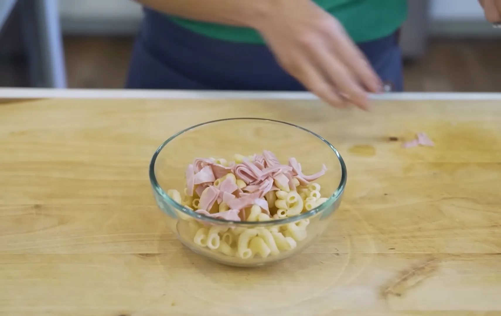 Adding sliced deli ham to cooked elbow macaroni in a glass bowl for cold pasta salad