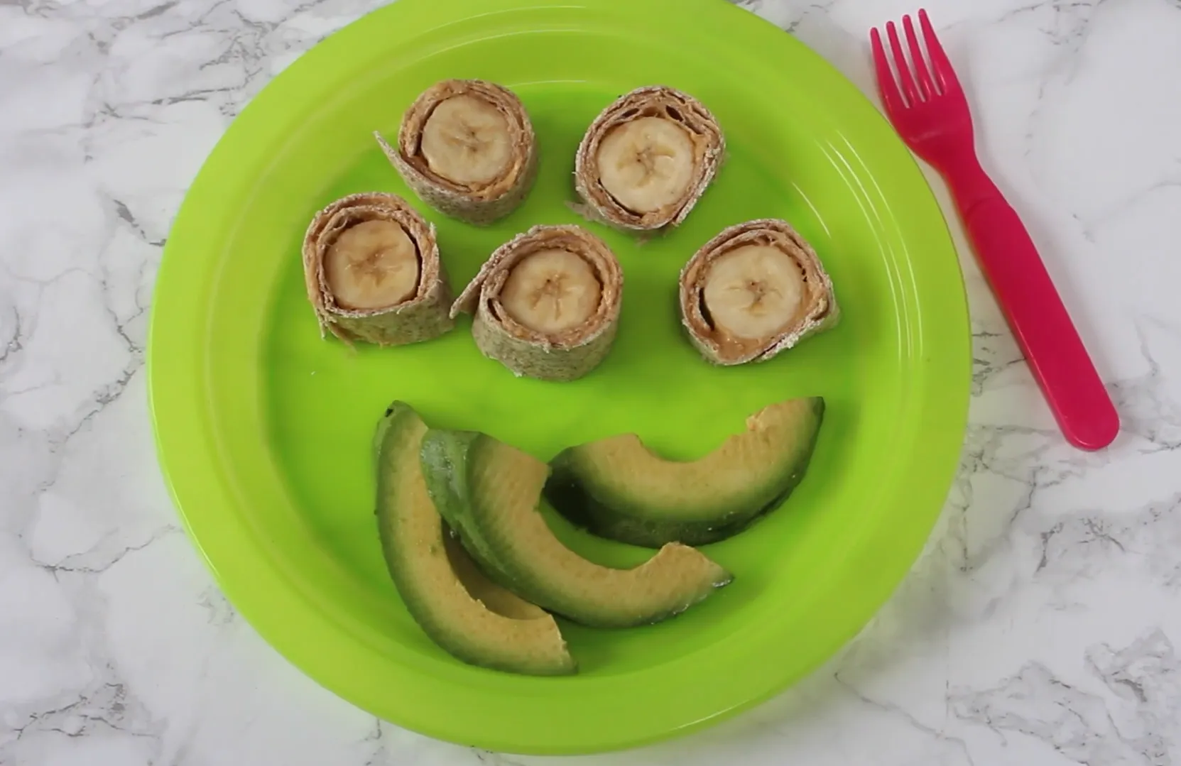 Banana and peanut butter tortilla roll-up pinwheels with sliced avocado arranged as a smile on a green plate