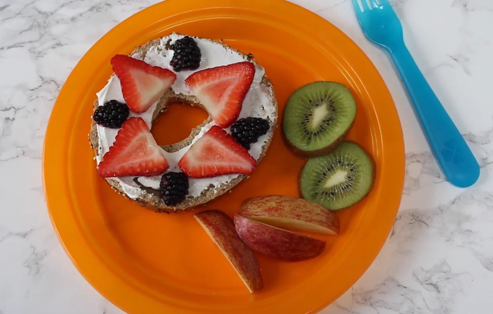 Cream cheese bagel half decorated with strawberries and blackberries in a star pattern alongside sliced kiwi and apples