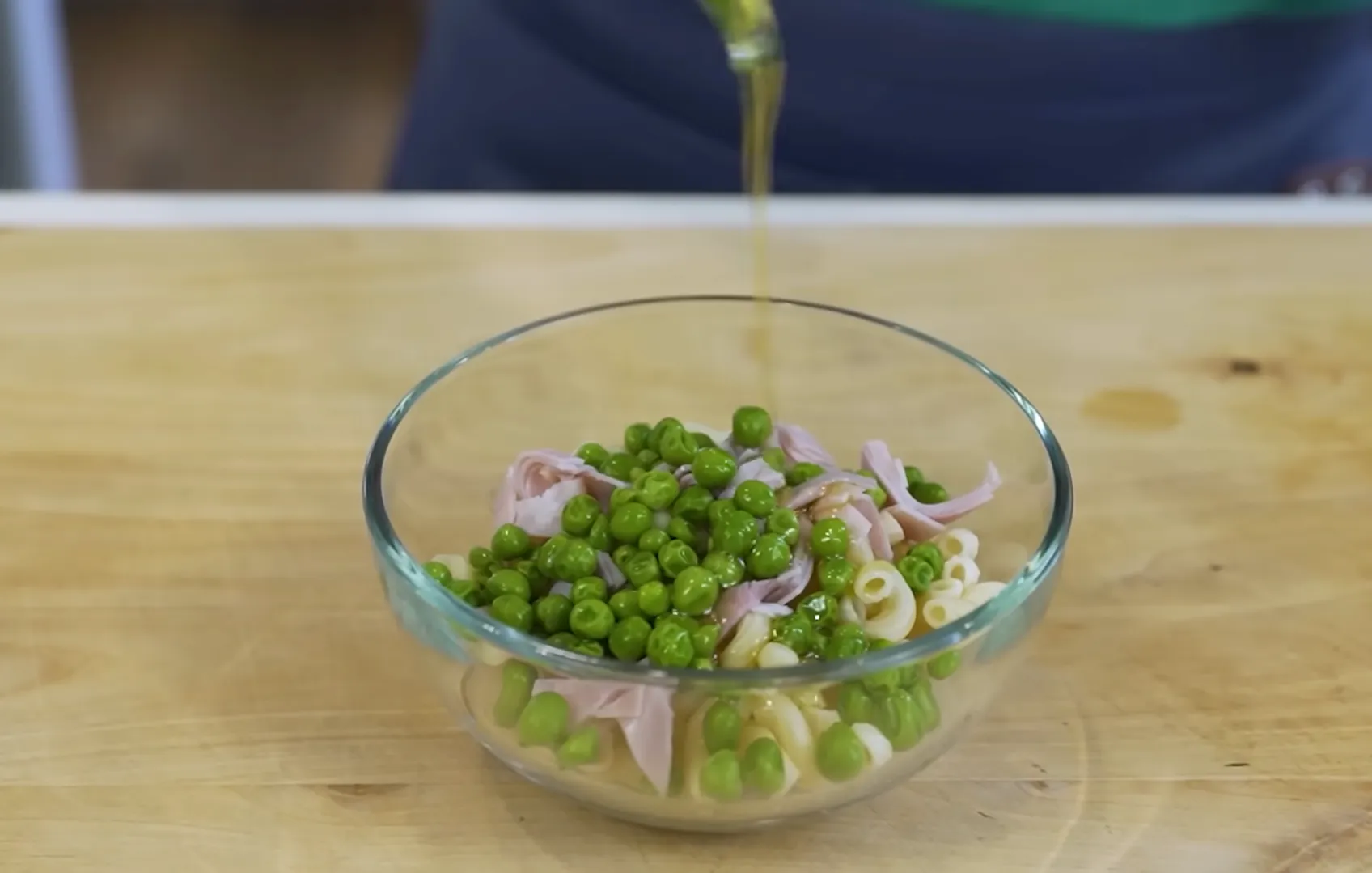 Drizzling olive oil dressing over cold macaroni, green peas, and ham pasta salad