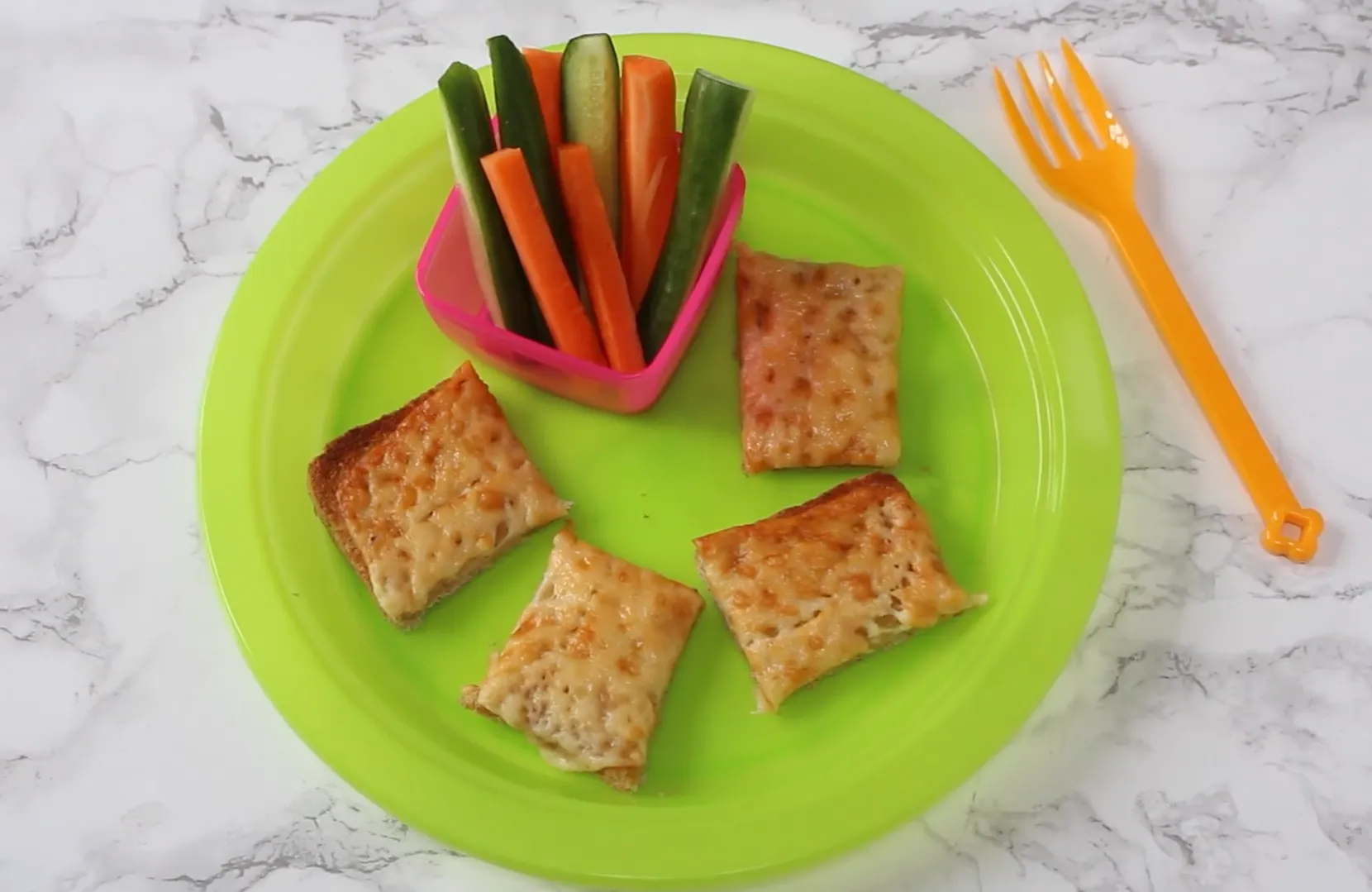 Melted cheese toast cut into squares with cucumber and carrot sticks on a bright green plate
