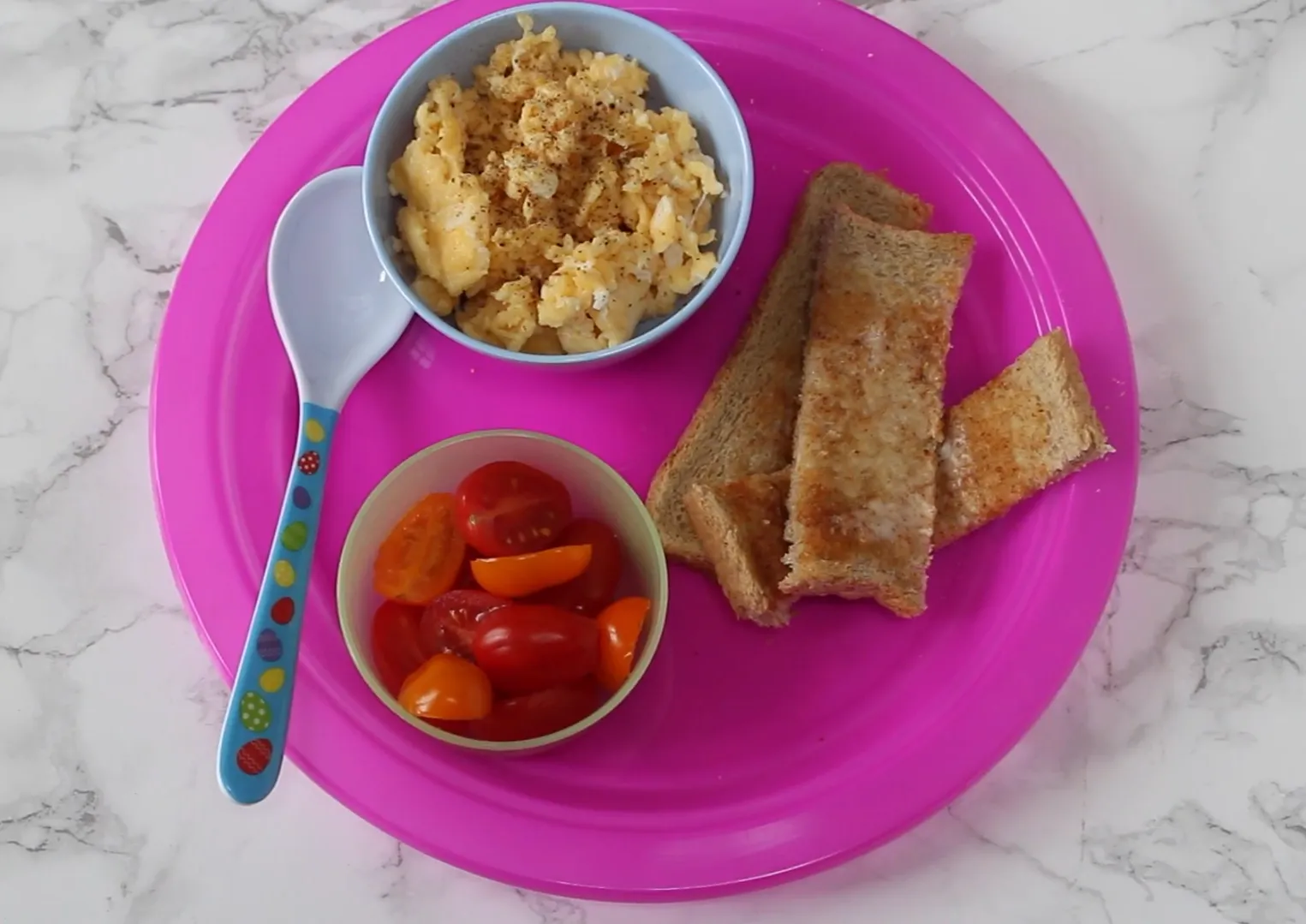 Soft scrambled eggs in a bowl with buttered toast soldiers and halved cherry tomatoes on a pink plate