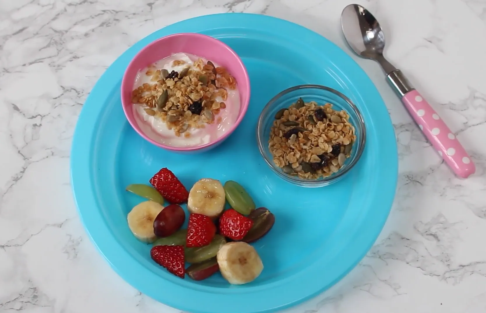 Yogurt bowl topped with granola served with sliced bananas, strawberries, and grapes on a blue plate