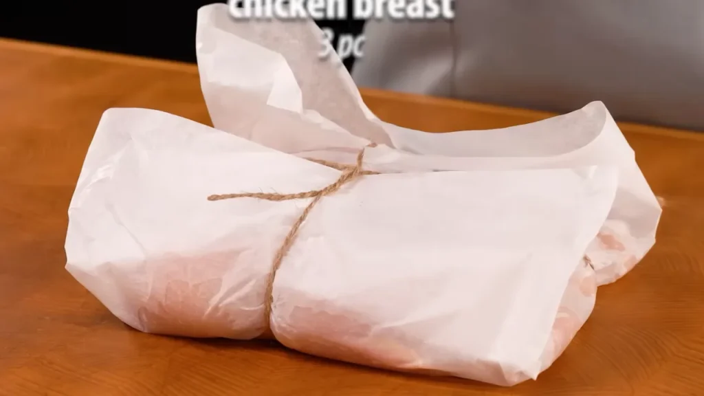 Chicken breast wrapped in white paper and tied with string on a wooden table, ready for pounding