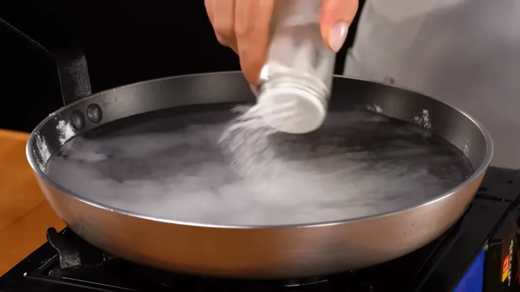 Adding salt from a shaker into a pan of water to prepare brine for chicken breast