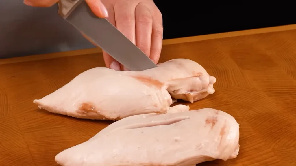 Cutting raw chicken breasts with a knife on a wooden cutting board before cooking